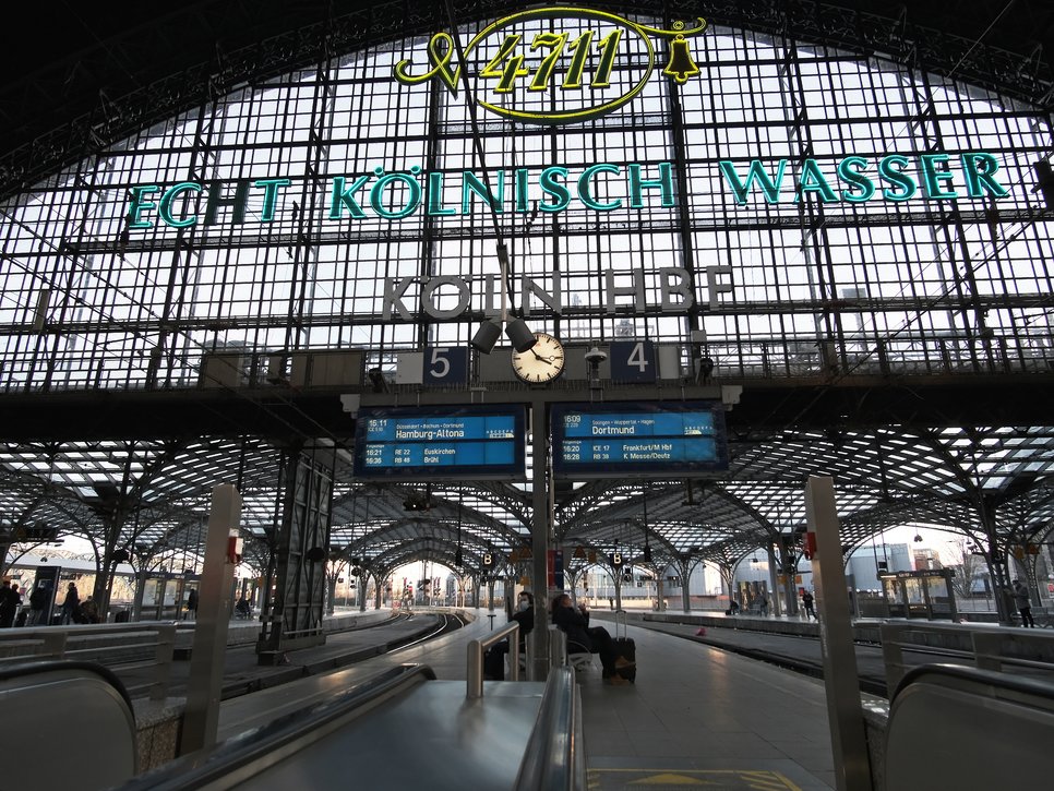 Ansicht des Kölner Hauptbahnhofs mit leeren Gleisen, einer zentralen Uhr und einem prominenten Echt Kölnisch Wasser-Werbeschild.
