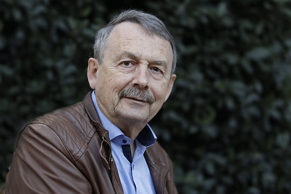 A person wearing a brown leather jacket and a blue shirt with a dark collar stands in front of a background of blurred foliage.