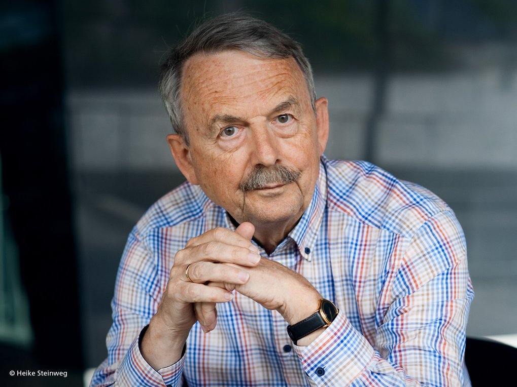 The photo shows a somewhat friendly-looking man wearing a blue, white, and red checkered shirt. He is sitting with his hands folded on a seat and staring at the camera with a questioning, slightly provocative look.