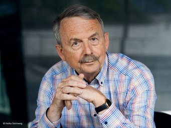 Wolfgang Streeck The photo shows a somewhat friendly-looking man wearing a blue, white, and red checkered shirt. He is sitting with his hands folded on a seat and staring at the camera with a questioning, slightly provocative look.