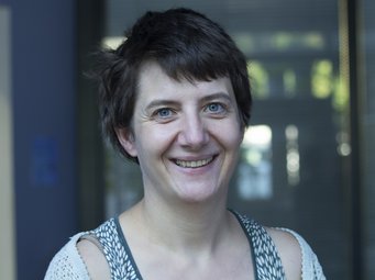 Helen Callaghan Eine Person, vermutlich weiblichen Geschlechts, mit kurzem, dunklem Haar ist in einem gemusterten Oberteil und einer Strickjacke zu sehen. Die Szene ist offenbar vor einer Glaswand lokalisiert, durch die diffuses Licht fällt, allerdings ist der Hintergrund stark verschwommen und hat keine eindeutigen Konturen. Die Person lächelt freundlich in die Welt, sie offenbart einen grundlegenden Optimismus, der ansteckend wirken könnte (Vorsicht).