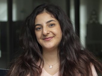 Ceren Çevik The photograph shows a specimen of the genus Homo sapiens, with external characteristics indicating that it is female. She has full, dark, wavy hair that falls forward over both shoulders and across her chest. Her dark brown eyes smile mischievously, and her head is tilted gently to the right. One cannot help but sense a deep love for humanity in this expression. The individual has a silver pendant necklace placed over the sternum, just above the beige top. In the background, numerous blurred windows can be glimpsed.