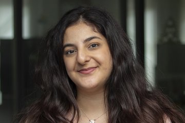 The photograph shows a specimen of the genus Homo sapiens, with external characteristics indicating that it is female. She has full, dark, wavy hair that falls forward over both shoulders and across her chest. Her dark brown eyes smile mischievously, and her head is tilted gently to the right. One cannot help but sense a deep love for humanity in this expression. The individual has a silver pendant necklace placed over the sternum, just above the beige top. In the background, numerous blurred windows can be glimpsed.