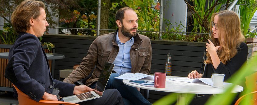 Three people are sitting at a round table outdoors and having a conversation. On the table are a laptop, documents, a bottle, and a cup. Plants can be seen in the background.